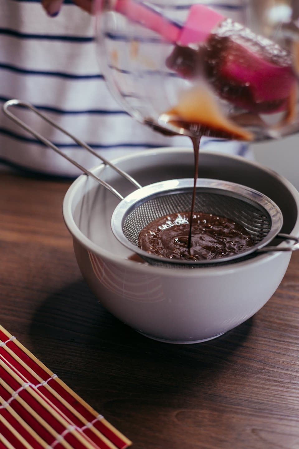 Hands pour chocolate mixture through a sieve into a bowl.